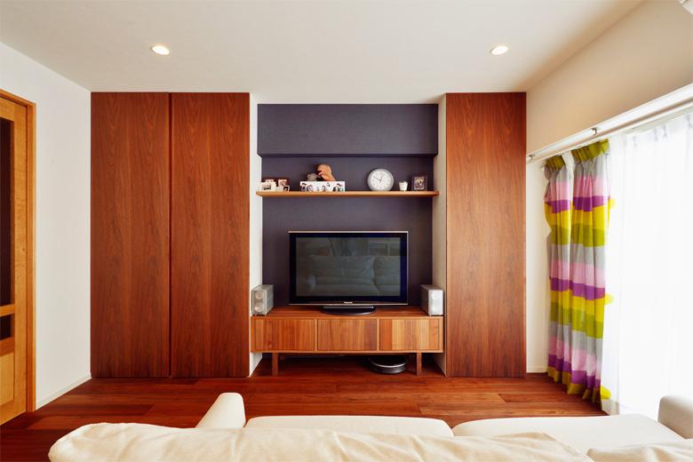 リビングのTVまわりには大容量の収納スペースを造作。左側にはリビングで使うものを収納。右側にはバルコニーやカーテン前のパイプに干した洗濯物を仕舞うことができて便利。扉は木目の美しいウォールナットの突き板。TV背面の壁は濃い紺色のアクセントクロスでモダンな印象に。