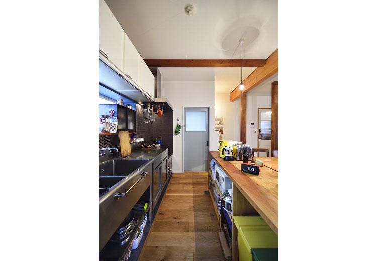 Mさんが中古で購入したステンレスの業務用キッチン。吊り戸棚は既存のものを再利用した。リビングとの間に置かれたカウンター収納は、Mさんのご両親が作ったオリジナル。奥に見えるグレーのドアも、Mさんが購入したもの。洗面室、浴室につながる