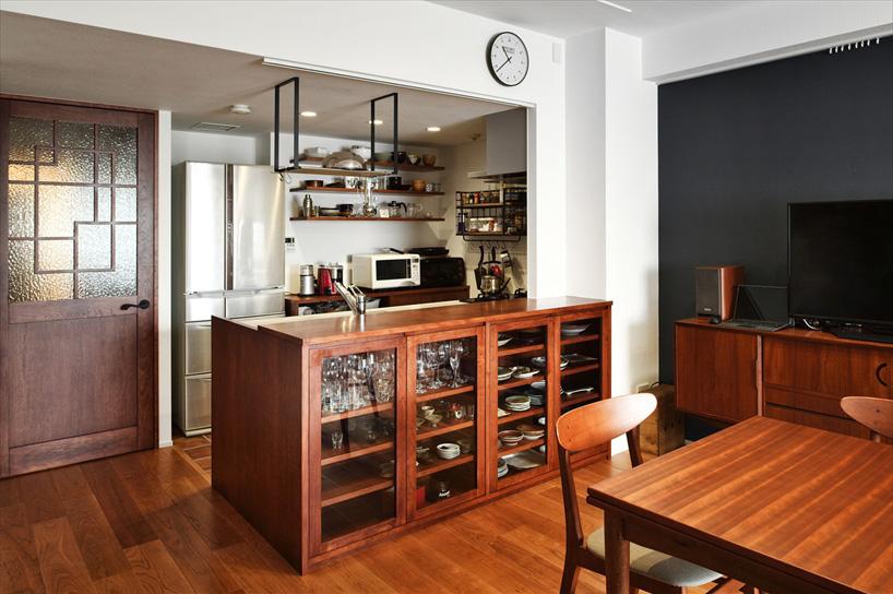 小窓が空いているだけで閉ざされた印象だったキッチンは、壁をなくしてオープンな対面キッチンに。カップボードを兼ねたキッチンカウンターを造作。お気に入りのグラスや食器が見えるよう、扉はガラスにした。シンクの上には、夫人のご希望によりステンレス製の水切り棚を。背面には家電収納棚を造作。