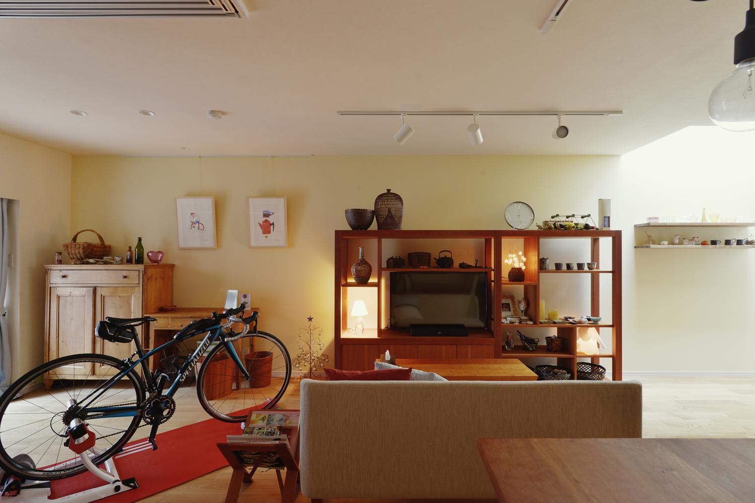 壁一面にイエローアイボリーの珪藻土を採用。Hさん愛用のチェリーの家具とも美しく調和する。テラス側にTVを見ながら自転車トレーニングをするスペースを確保。TVボードの右側にはコレクションを並べる飾り棚を設置した