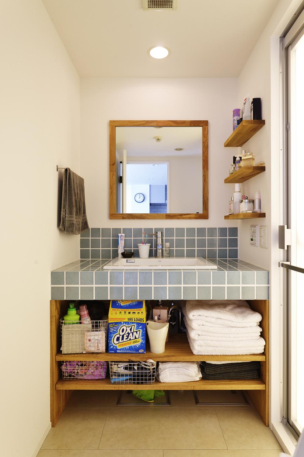 造作の洗面台はブルーグレイのタイル貼りに白い目地で爽やかな印象に仕上げた。下部のオープン収納やサイドの棚、鏡の枠まで同色の木を用いている点にもご夫妻のこだわりがたっぷり詰め込まれている