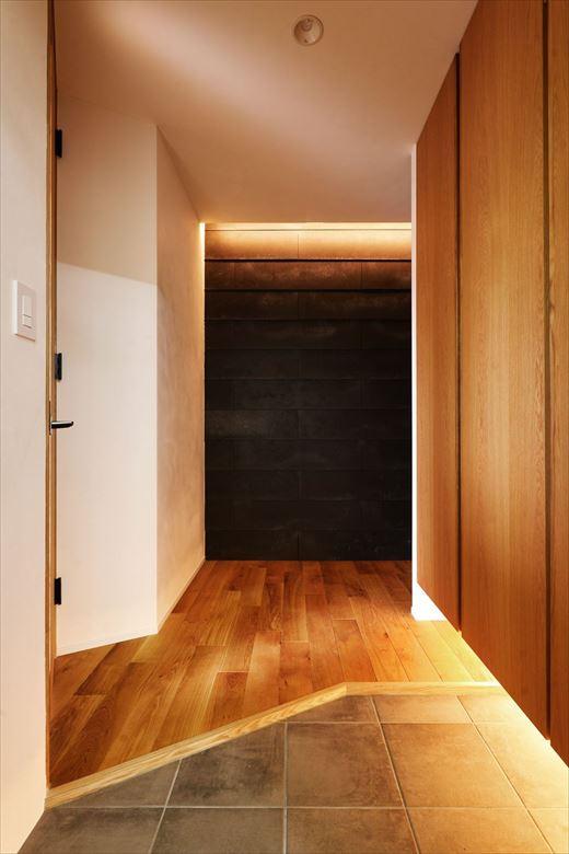リビングの板壁と同シリーズの外壁材を用いた玄関ホール。テレビ背面がフラットなのに対し、こちらは鎧貼りを採用。収納は天井までの大容量に変更し、スッキリとしたデザインと間接照明で空間の広がりを感じることができる