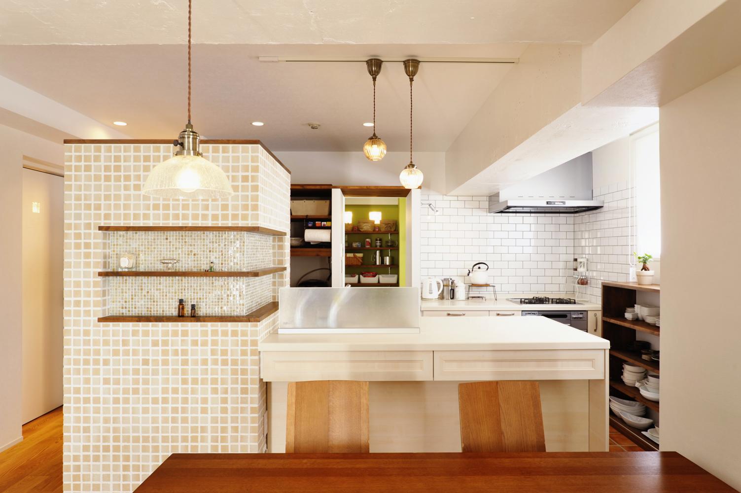 リビングを向いたオープンなカウンターキッチン。カウンター横の壁は冷蔵庫の目隠し。天井との間に空間をつくり、明るいタイルと棚部分にはガラスモザイクを入れ、圧迫感を感じさせないデザインにした。キッチンの奥に見える明るいグリーンのクロスはパントリー