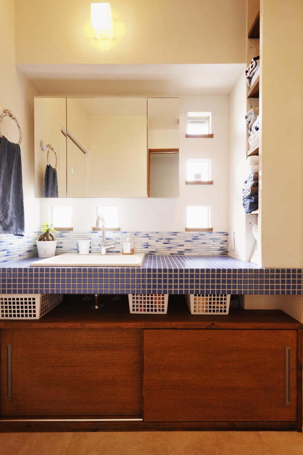 洗面カウンターと壁には、Mさんが好きなブルーのタイルを２種類組み合わせて配置。カウンター下の収納はタモ材で造作。カウンター横の壁にも収納棚を設け、洗面室で使うタオルなどは使う場所に収納できるようにした。