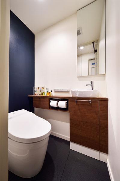 濃紺を一面に配色した落ち着いた装いのトイレです。壁に沿って腰高の収納棚を設置しました。幅10数センチの棚上に手洗いボウルも設置。奥行が浅い棚収納は圧迫感を与えません。鏡の裏にも収納が隠されています。
