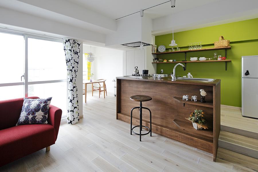 明るい陽の光がたっぷりと降り注ぐLDK。壁の鮮やかなグリーン、温もりある無垢材のキッチンカウンターのブラウン、ソファの赤い配色が、女性らしい華やかな空間を演出。洋室（写真中央奥）の間仕切りを取り払うことで、陽の光や風が室内のすみずみまで行き渡り、気持ちのよい空間に生まれ変わりました。