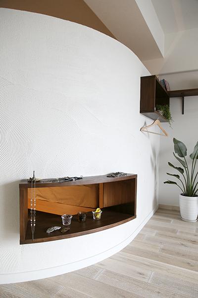 Rの曲線が優雅な壁は、LDKと寝室を優しく区切るとともに、空間のアクセントにも。壁に創った通風・採光用の開口部には棚を造作し、雑貨や小物を飾るスペースに。写真奥の高い位置に設けた飾り棚には溝を入れ、ハンガーが掛けられるように工夫。ゲストの上着やお気に入りの洋服を掛ければ、まるでショップのような雰囲気