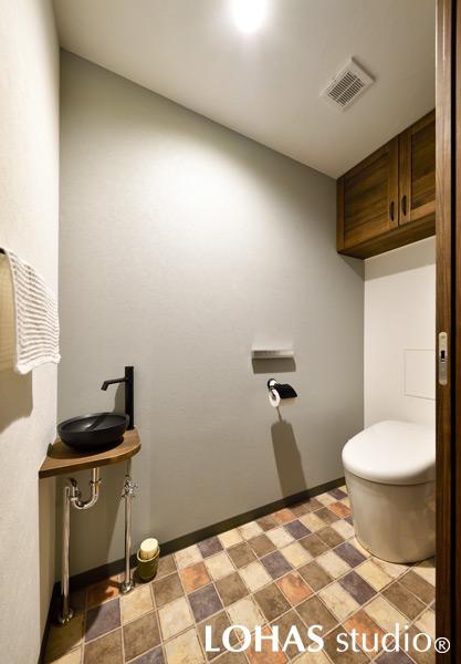 アンティークタイル柄のクッションフロアが優しい印象を与えるトイレです。代わりに、紙巻器や手洗いボウルはシックなブラックで引き締めました。吊戸棚を造作して収納力をアップさせています。