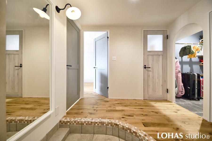 アール壁を使って柔らかく優しい印象となった玄関スペースです。大小のタイルを組み合わせて可愛らしくまとめ、大きな姿見でお出かけ前の身支度もばっちり。上がって右手にはスーツケースやゴルフ用具もまとめて収納できるシューズクロークをお作りしました。