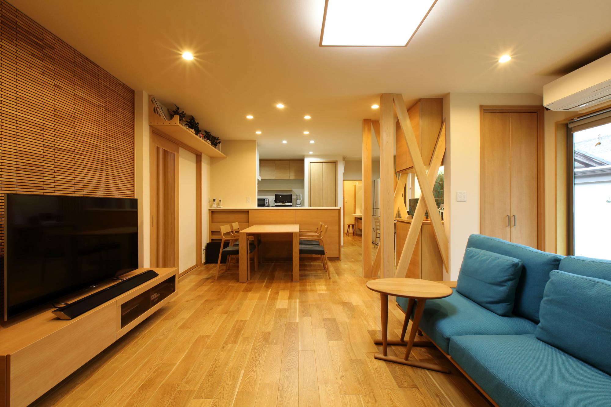 木質感あふれるLDKは、和室や廊下などを繋げてひとつの空間に。明るいオークのフロアには床暖房を設置。TV背面の壁面は当社オリジナルのウッドタイルで仕上げ、室内の引き戸には縦格子のガラス戸を採用。出入り口の上部には立派な神棚を設けた。