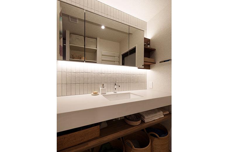 洗面ボウルの両脇にカウンターのある広い洗面台を建築士が提案。広くする必要があるかなと思っていたそうだが、実際生活してみると、朝の時間夫婦で同時に使ってもストレスがなく動けて、物はいっぱい入るので採用して良かったとか。壁のタイルに間接照明で落ち着く空間にした。