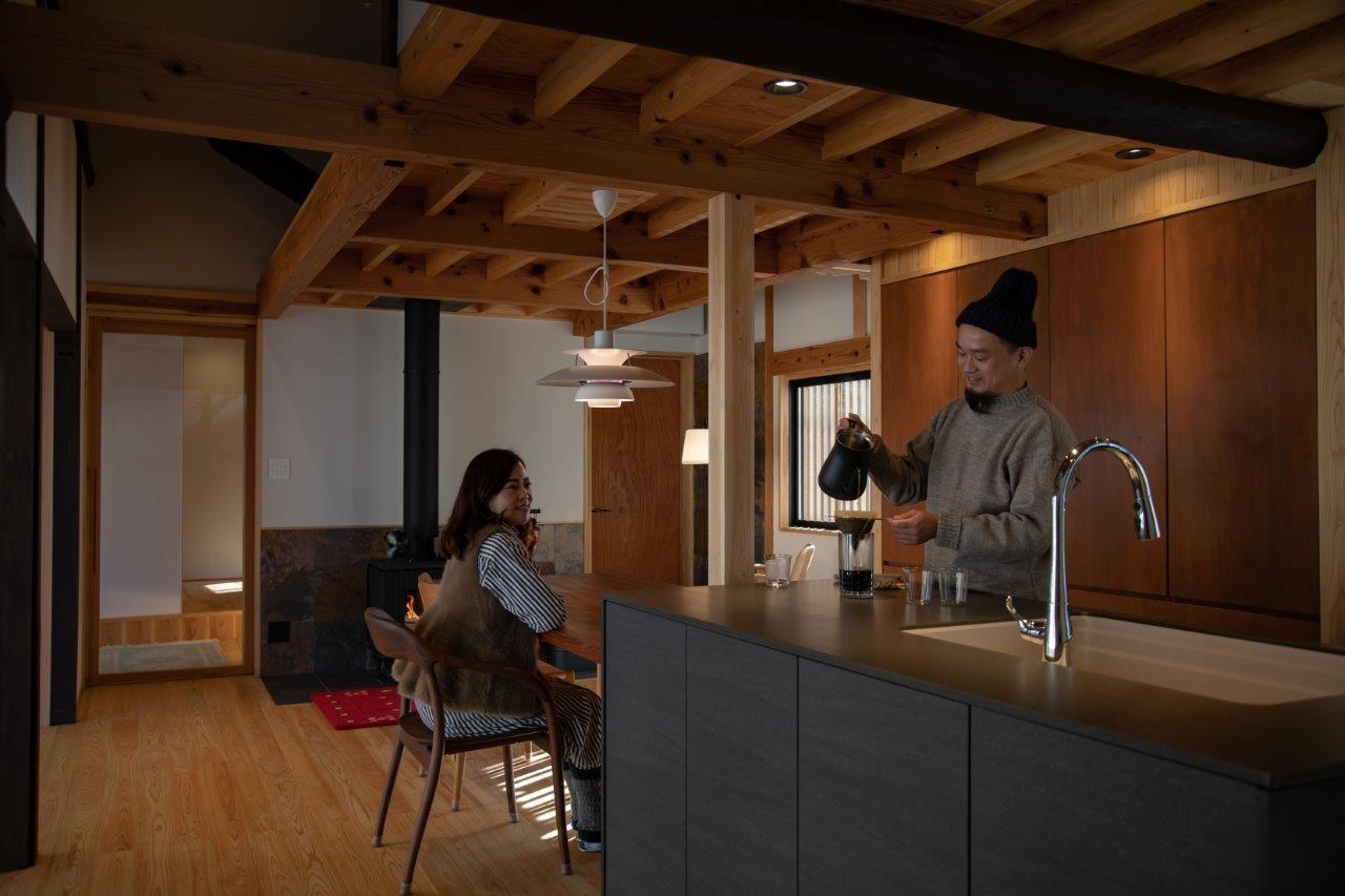 木の温もりを存分に活かした、落ち着きのある住空間へと生まれ変わったリフォーム事例です。天井や梁、床に無垢材をふんだんに使用することで、自然素材ならではのやさしい雰囲気を演出しています。

キッチンはオープンスタイルに刷新され、家族や来客との会話を楽しみながら過ごせる設計に。カウンター越しにコミュニケーションが生まれる、暮らしの中心となる空間です。モダンな設備と木の質感が絶妙に調和し、機能性とデザイン性を両立しています。

また、奥に見える薪ストーブが空間全体にぬくもりをもたらし、冬でも心地よく過ごせる工夫が施されています。照明もやわらかな光を選び、リラックスできる空気感を引き立てています。