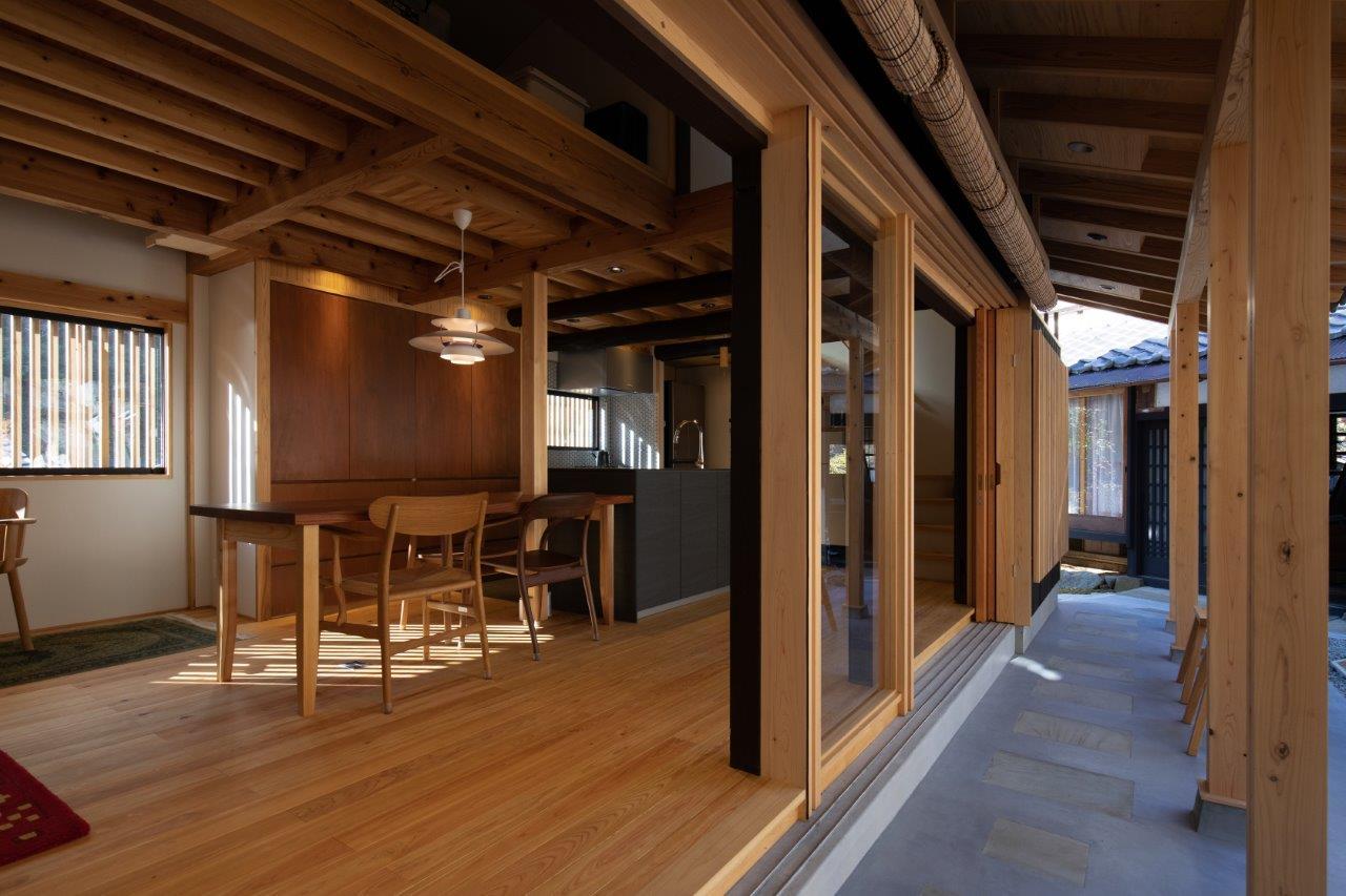 内と外をゆるやかにつなぐ、開放感あふれるリフォーム事例です。無垢材の床と天井が連続することで、室内にいながらも外の空気を感じられるような心地よい空間が広がっています。

大きく開かれた開口部からは自然光がたっぷりと入り、ダイニングスペースを明るく包み込みます。外部の土間空間とフラットにつながる設計により、季節や時間の移ろいを日常の中で楽しめるのが魅力です。

また、奥に見えるキッチンとの一体感も美しく、生活動線にも配慮された機能的なレイアウトとなっています。木の温もりとシンプルなデザインが調和し、長く愛される住まいへと生まれ変わりました。