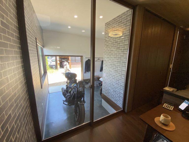 居室とバイクガレージを大きなガラス建具で仕切り、室内から愛車を眺められる空間としました。

ガラス越しに視線が抜けることで、居室とガレージに一体感を持たせながら、それぞれの空間を独立して利用できます。

レンガ調の壁とダークブラウンの木質仕上げを組み合わせ、落ち着いた雰囲気に。

日常のくつろぎの中